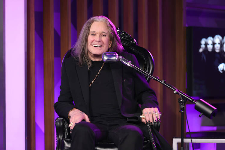Ozzy Osbourne smiles while sitting in chair in black outfit