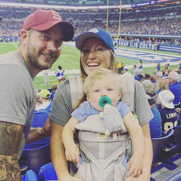 Mina Starsiak Hawk pses next to husband Steve Hawk and baby Jack at baseball game