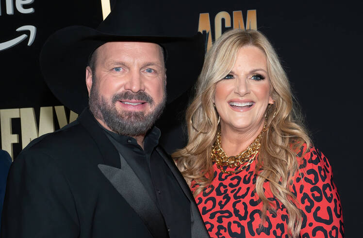 Garth Brooks, wearing an all black suit, poses next to wife Trisha Yearwood who's wearing a red leopard print dress.